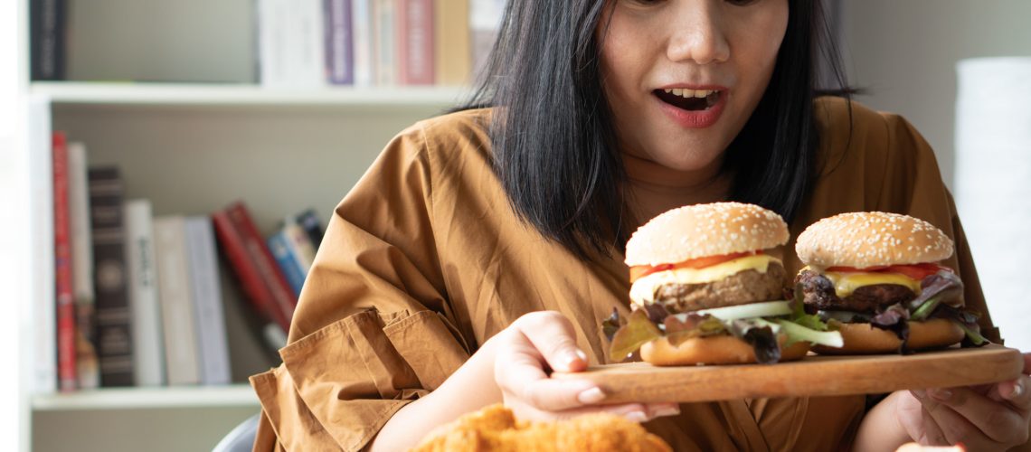 Hungry overweight woman holding hamburger on a wooden plate, Fried Chicken and Pizza on table, During work from home, gain weight problem. Concept of binge eating disorder (BED).